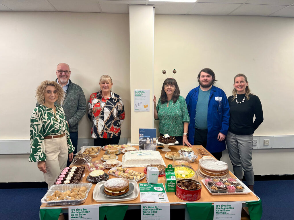 A group of employees at the Hayward Tyler Luton site stand behind a table filled with a variety of homemade and donated cakes for a charity bake sale. The event was held in support of Macmillan Cancer Support, with staff contributing by baking or donating sweet treats. The table is filled with an assortment of cakes, cupcakes, and pastries, with signs indicating the charity event. The team is smiling as they pose for the photo in an office setting, celebrating their successful fundraising effort, which raised £760 for the cause.